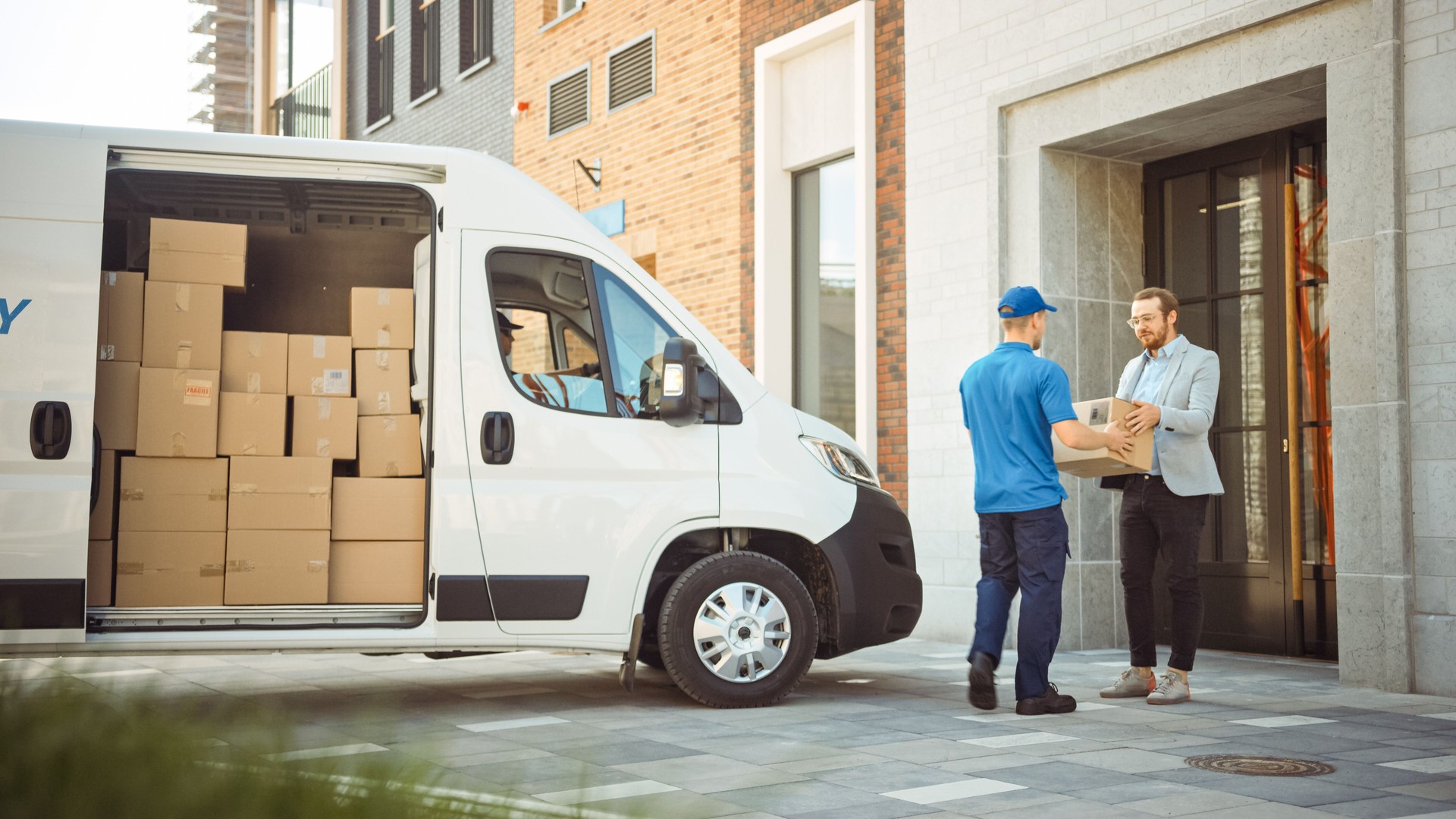 Delivery Man entrega un paquete postal a un cliente de negocios, que firma un dispositivo POD de firma electrónica. En el elegante área de oficina urbana moderna Courier entrega paquete de caja de cartón a un hombre